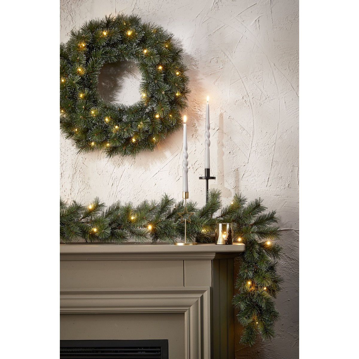 Christmas wreath and garland with lights on a mantelpiece, two white candles lit.