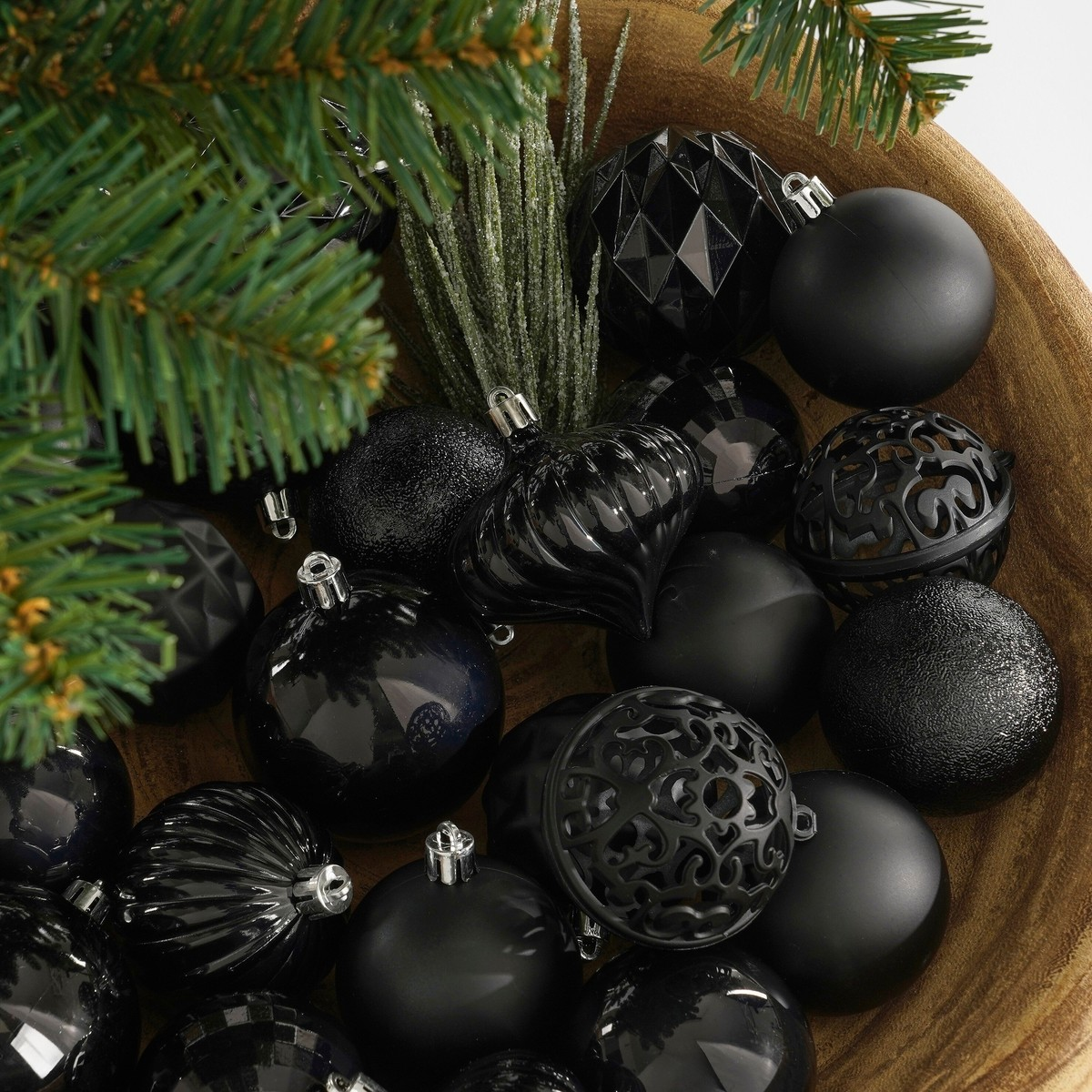 A wooden bowl filled with matte and shiny black Christmas ornaments and pine branches.