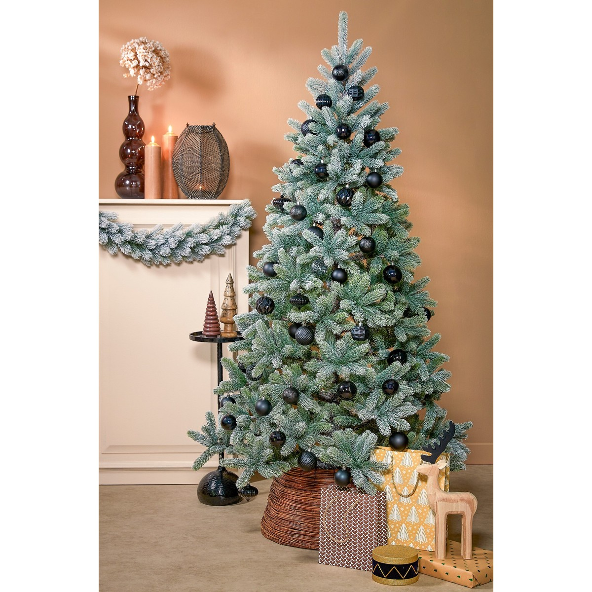 An artificial, lightly snow-dusted Christmas tree is decorated with black baubles. Gifts and decor sit nearby.