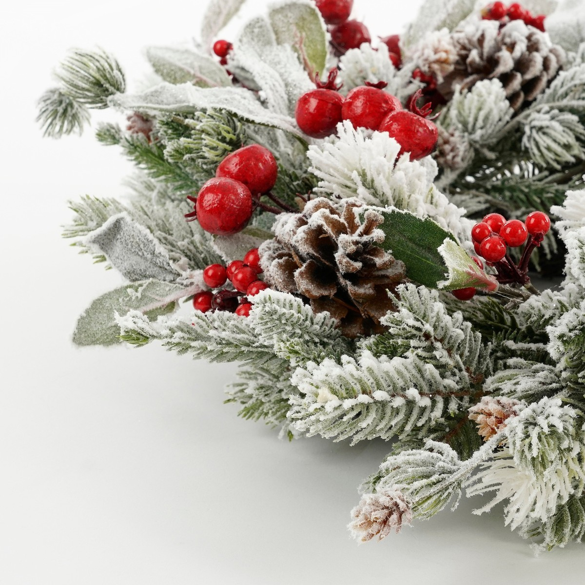 A snowy holiday wreath featuring green pine branches, red berries, and brown pinecones against a white background.