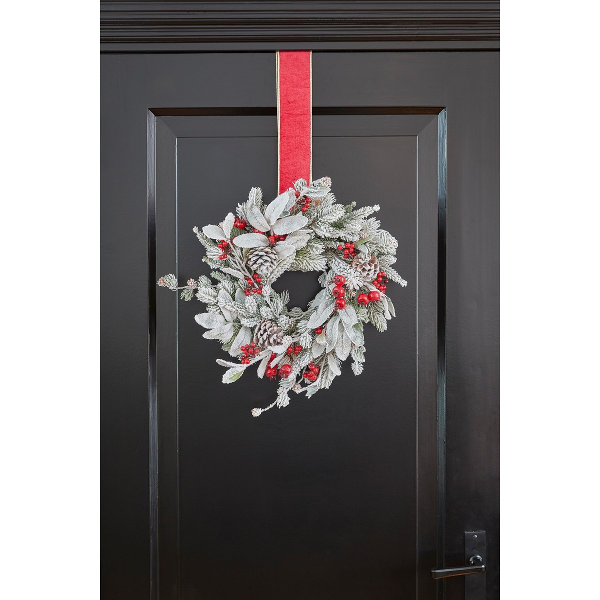 A frosted Christmas wreath with red berries and pinecones hangs by a red ribbon on a black door.