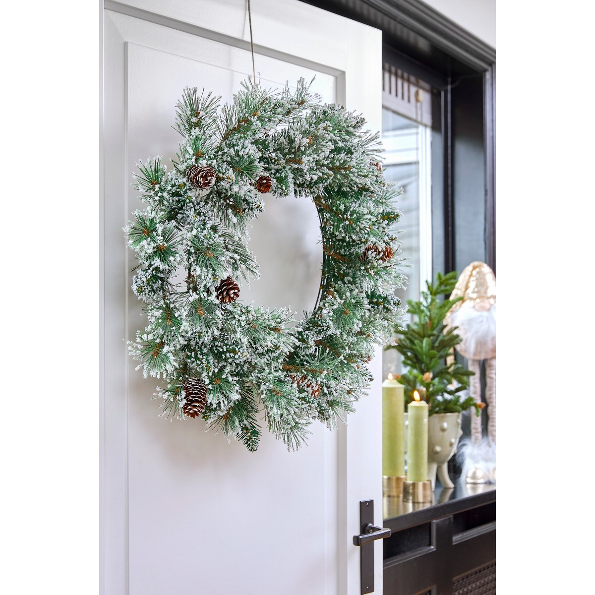 A frosted pine wreath with pinecones hangs on a white door, with candles in the background.