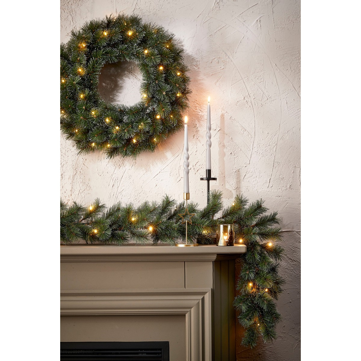 A lit Christmas wreath and garland adorn a fireplace mantel with two tall white candles against a textured wall.