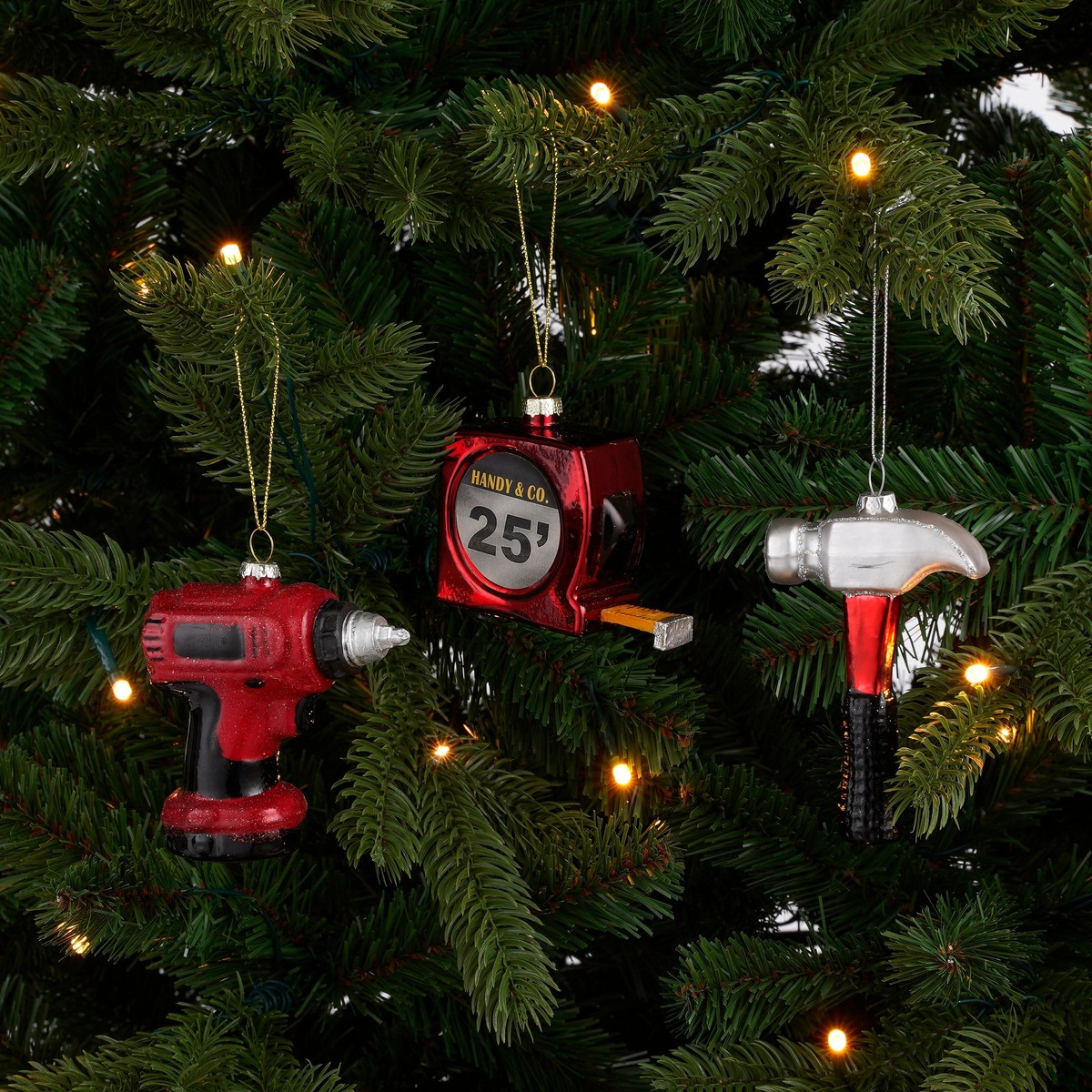Three tool-shaped Christmas ornaments: a red drill, a tape measure, and a hammer hanging on a lit tree.