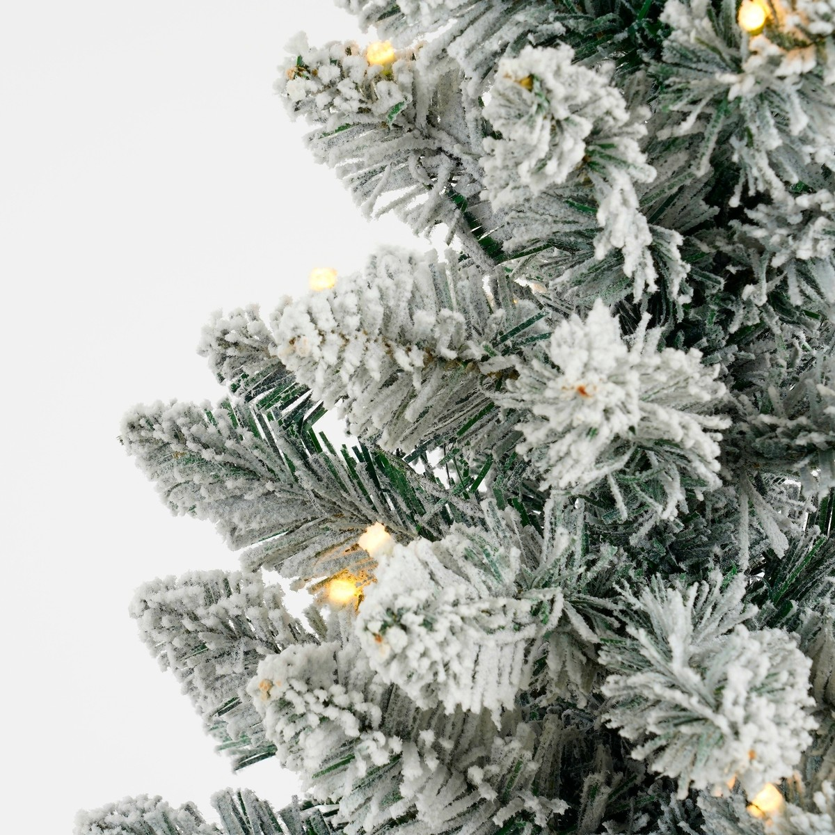 Close-up of artificial snow-flocked Christmas tree branches with small warm lights against a white background.