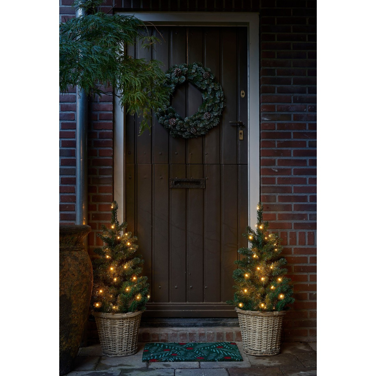 A dark brown wooden door is decorated with a pine wreath. Two illuminated pine trees in baskets stand in front of it.