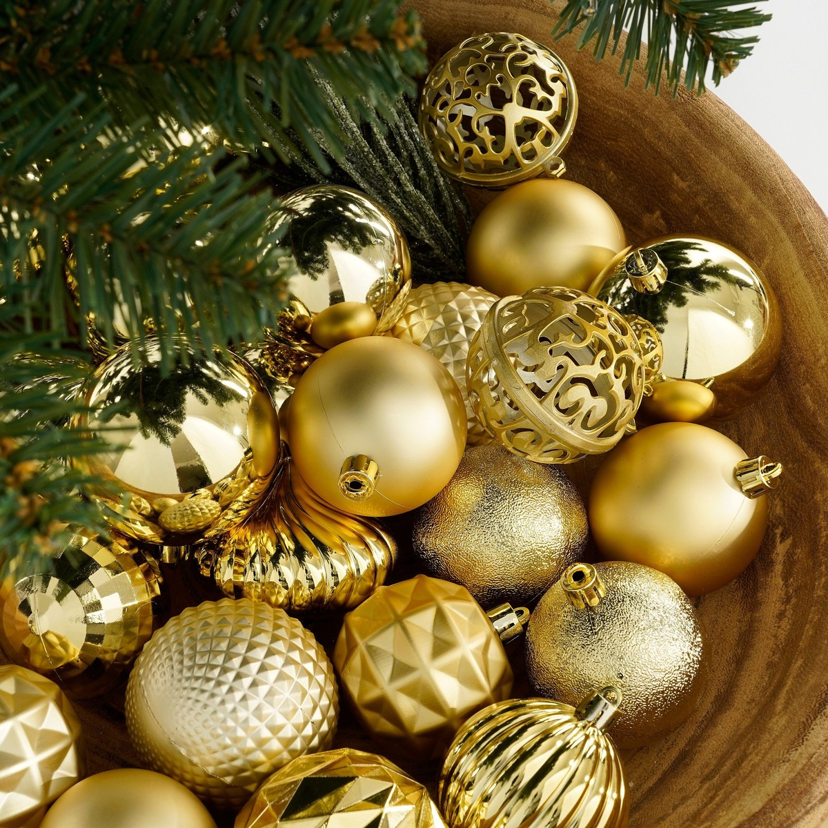 A wooden bowl filled with various shiny gold Christmas ornaments and pine tree branches.