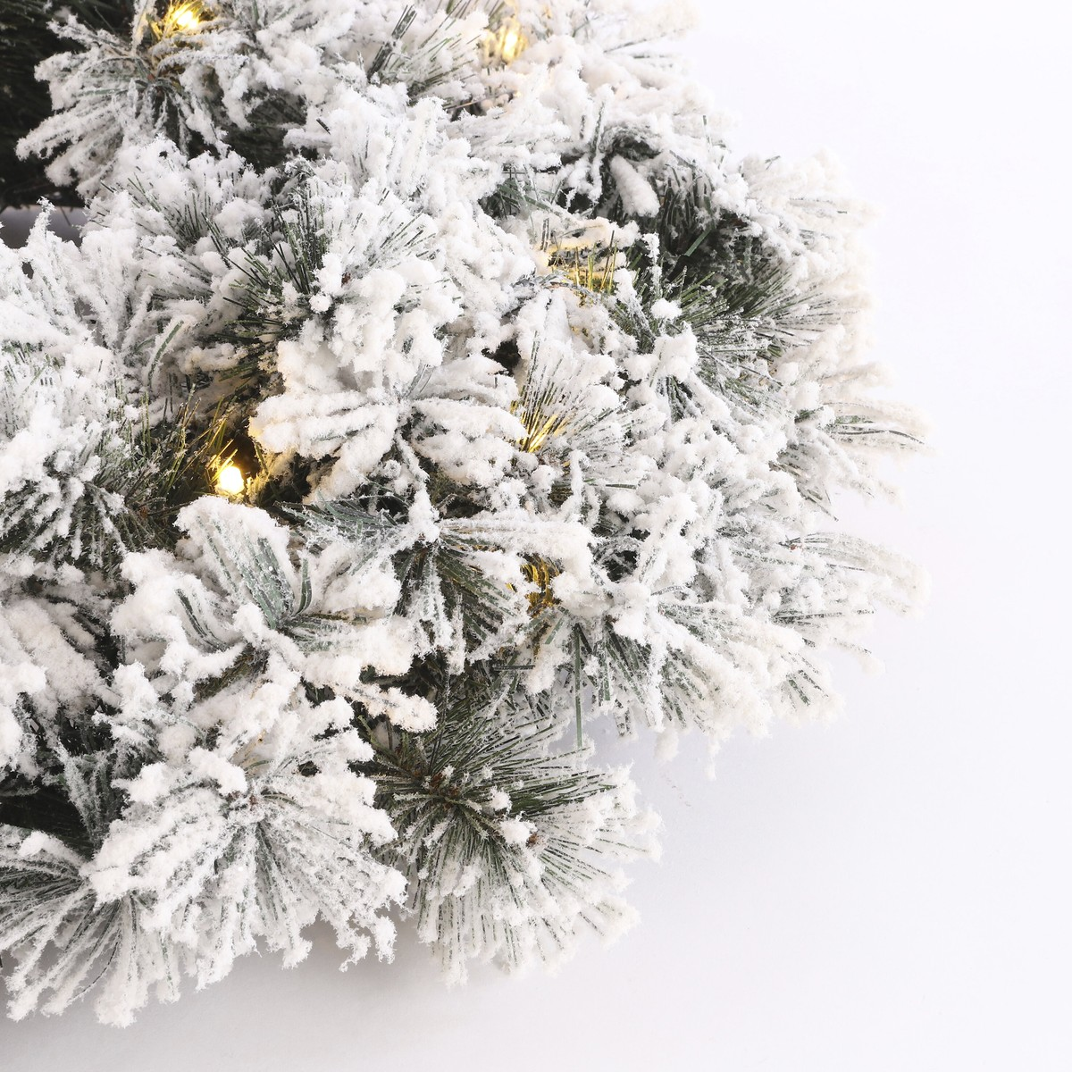 An artificial pine wreath covered in fake snow with warm lights against a white background.