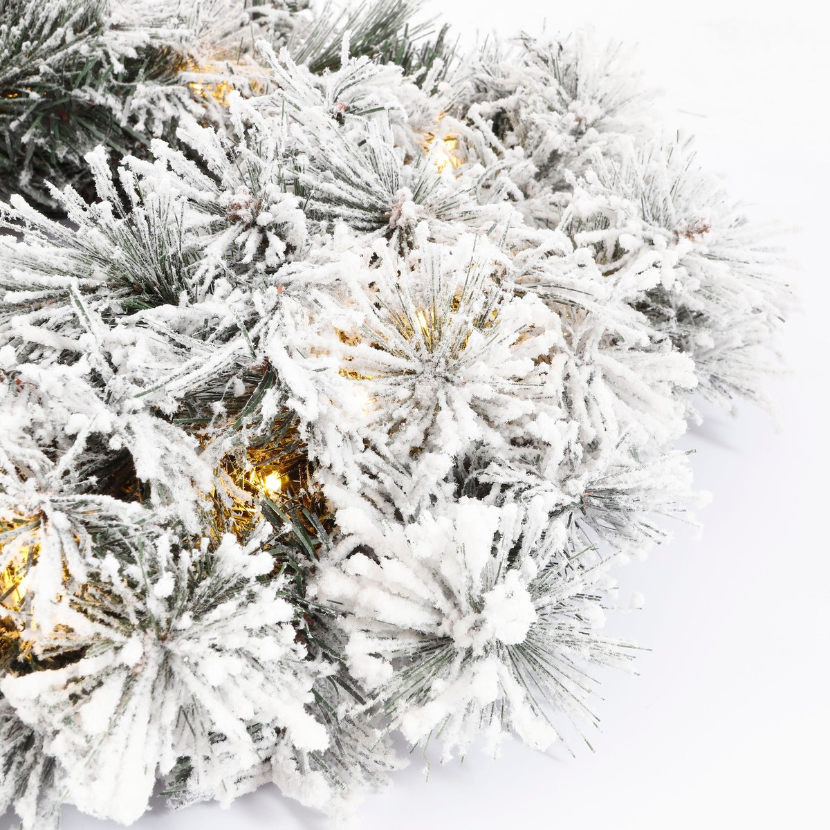 A snowy artificial pine wreath with warm lights against a white background.