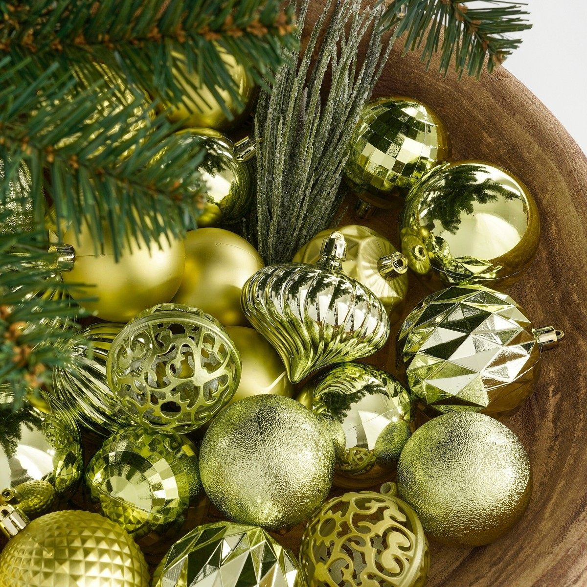 Gold and green Christmas ornaments and pine branches fill a wooden bowl.