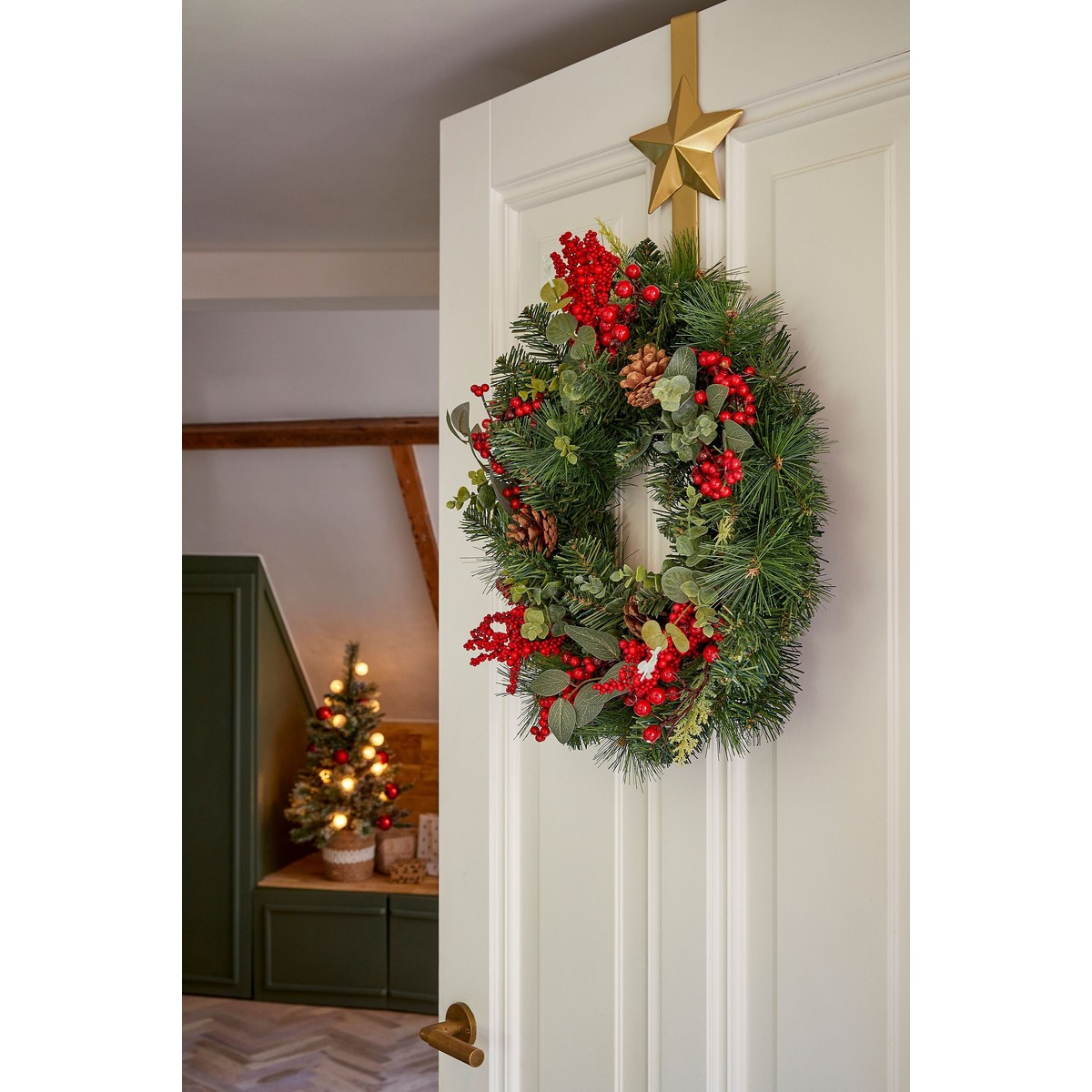 A Christmas wreath with red berries and pinecones hangs on a white door beneath a golden star.
