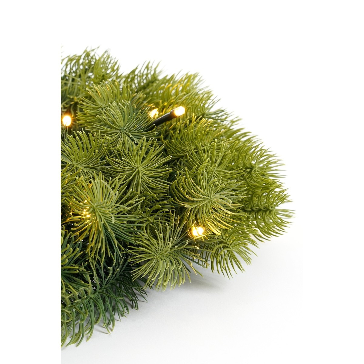Close-up of artificial green pine branches intertwined with small, warm white fairy lights on a white background.