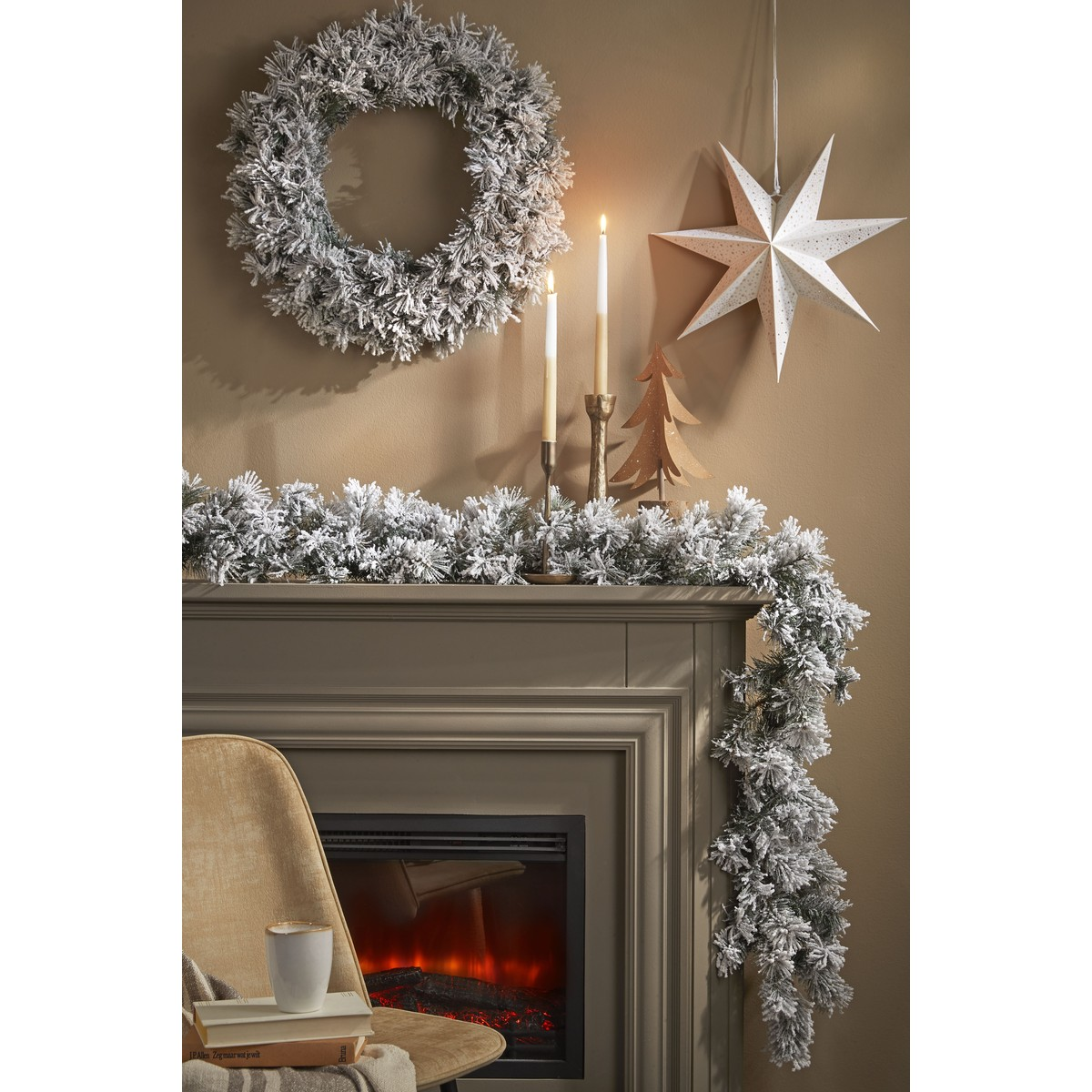 Fireplace decorated with snowy garland, wreath, lit candles, hanging star, and a chair with a mug.