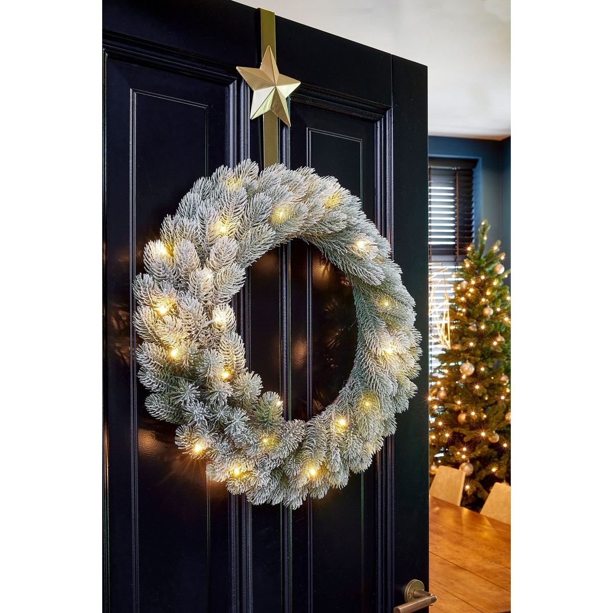 A frosted, lighted Christmas wreath with a gold star hangs on a black door.