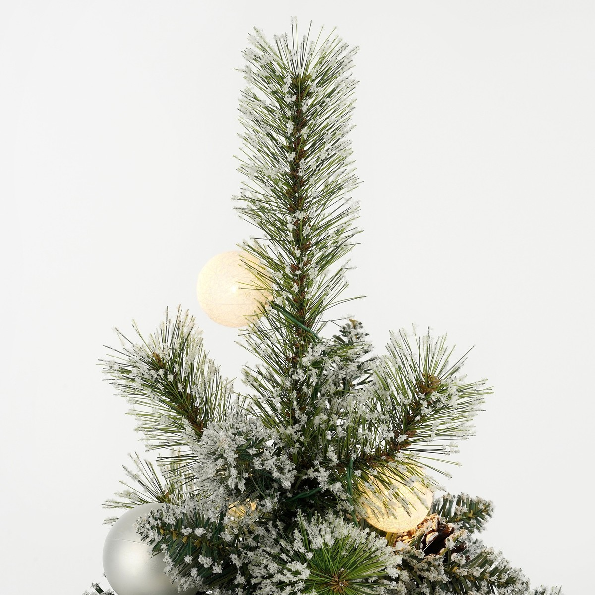 Snowy Christmas tree with lights and a silver ornament against a bright background.