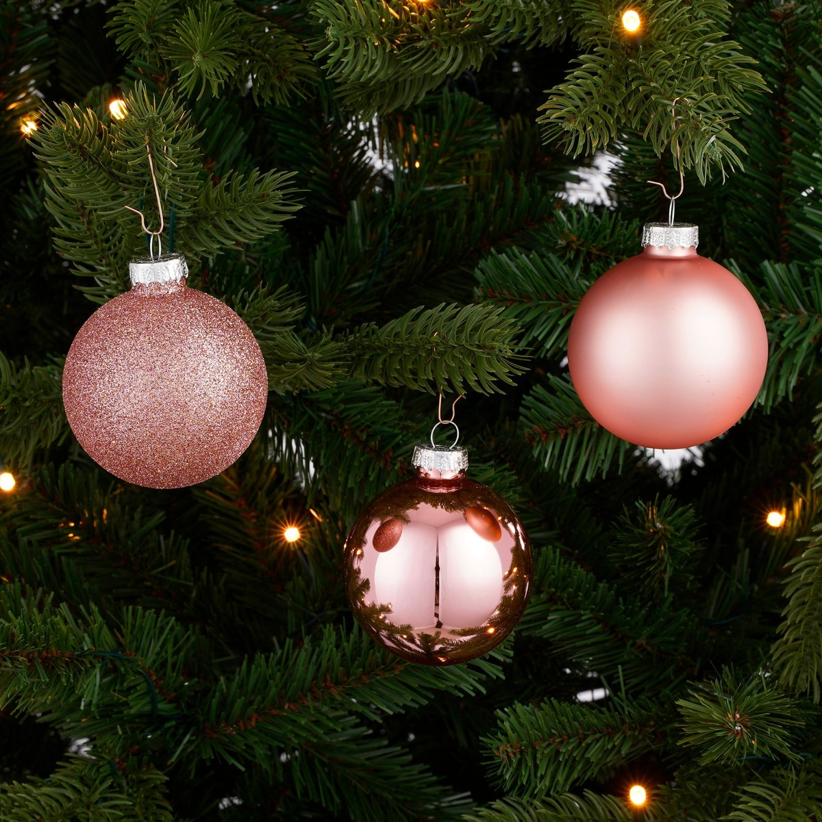 Three pink Christmas baubles in different finishes hang on the branches of a fir tree with small lights.