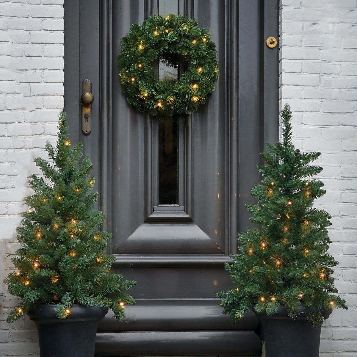 Two small lit Christmas trees in pots flanking a dark front door, with a lit wreath hanging above.