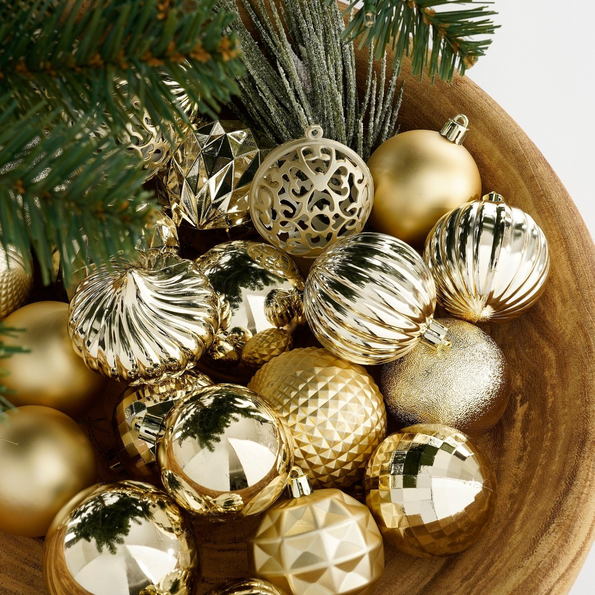 A wooden bowl filled with various gold Christmas ornaments and pine branches.
