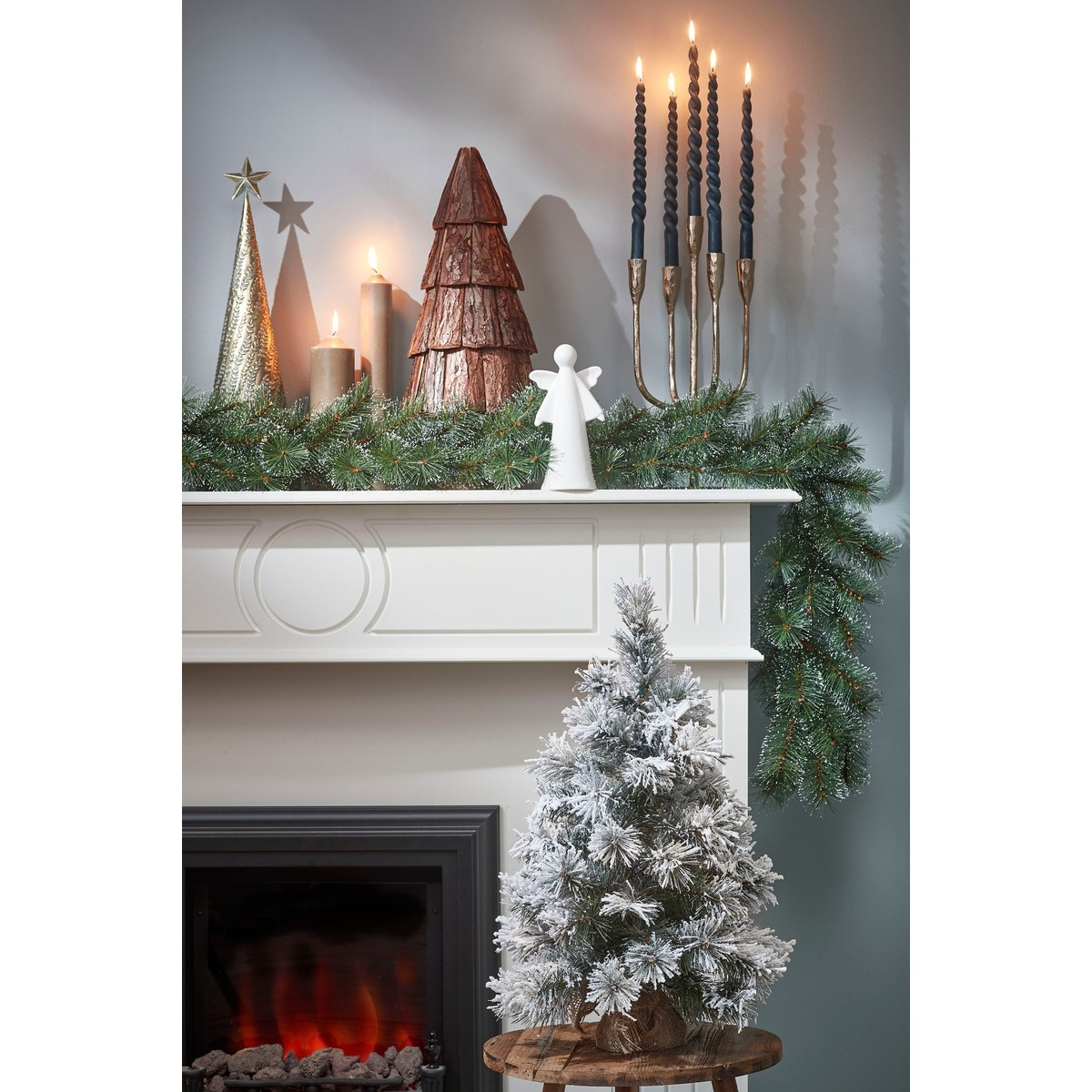 Festive mantelpiece with pine garland, lit candles, wood and metal trees, and a white angel figurine.