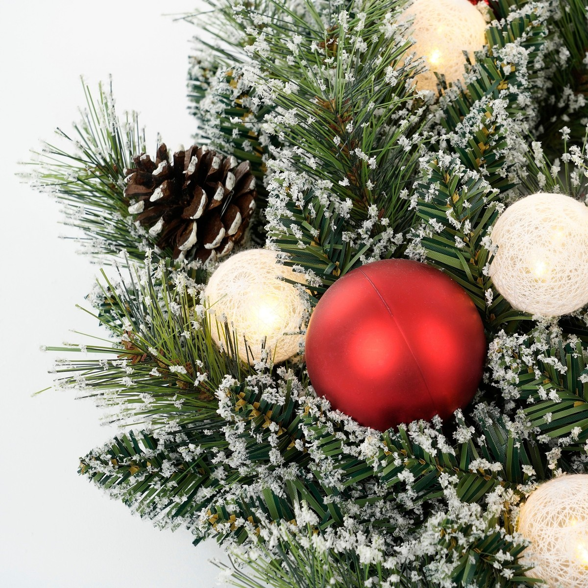 Snow-dusted pine branches decorated with a red ornament, a pinecone, and glowing white string lights.