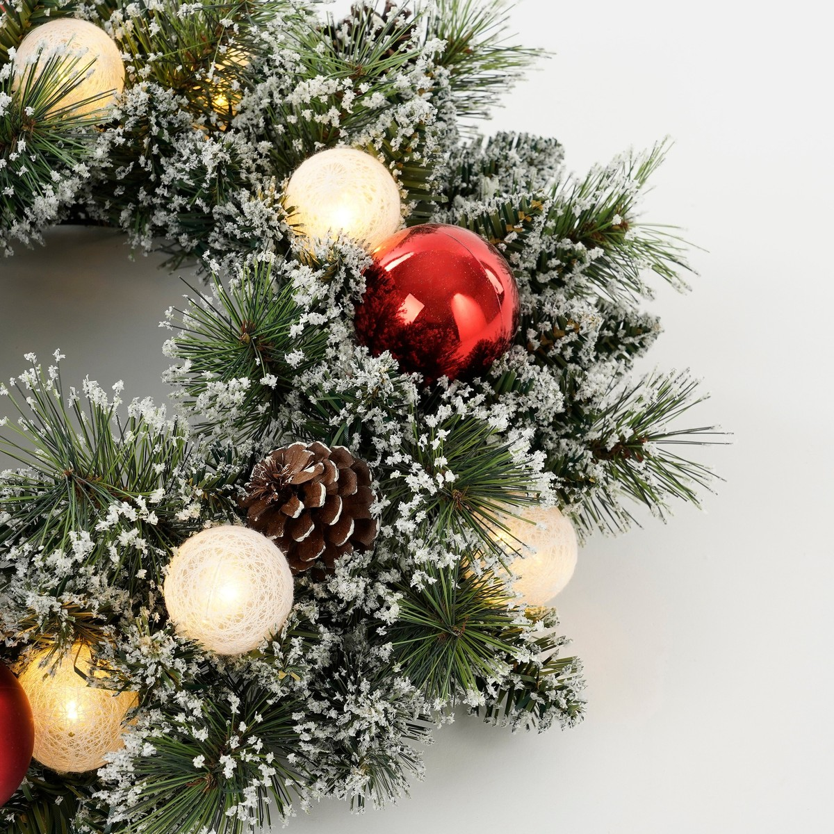 Snowy Christmas wreath with pinecones, red ornaments, and warm glowing string lights on a white background.