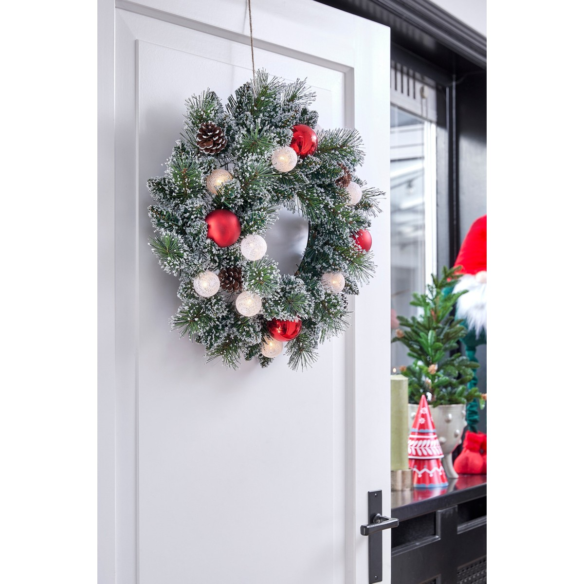 A snowy Christmas wreath with red ornaments and lights hangs on a white door.