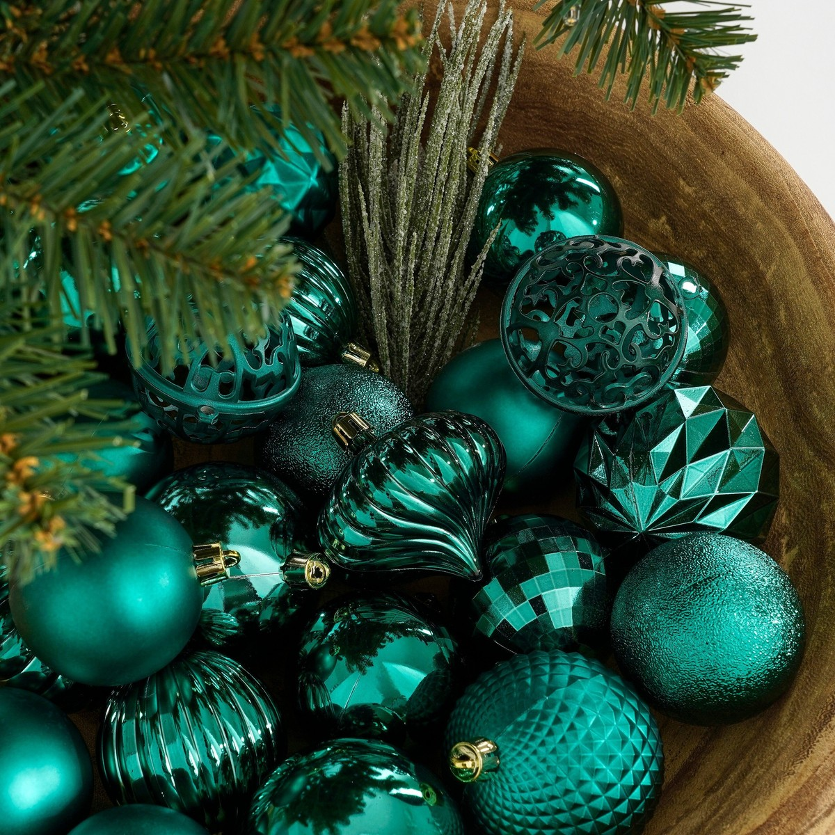 A wooden bowl filled with various shiny and matte turquoise Christmas baubles next to fir branches.