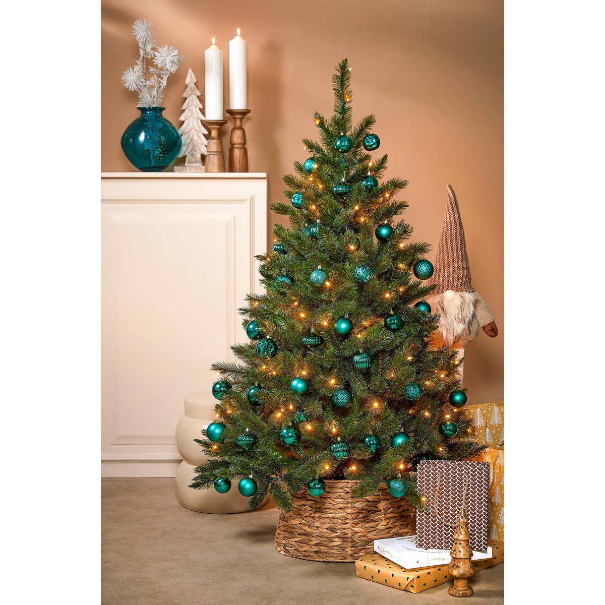 A decorated Christmas tree with teal ornaments and warm lights stands in a basket, with gifts and decor in the background.