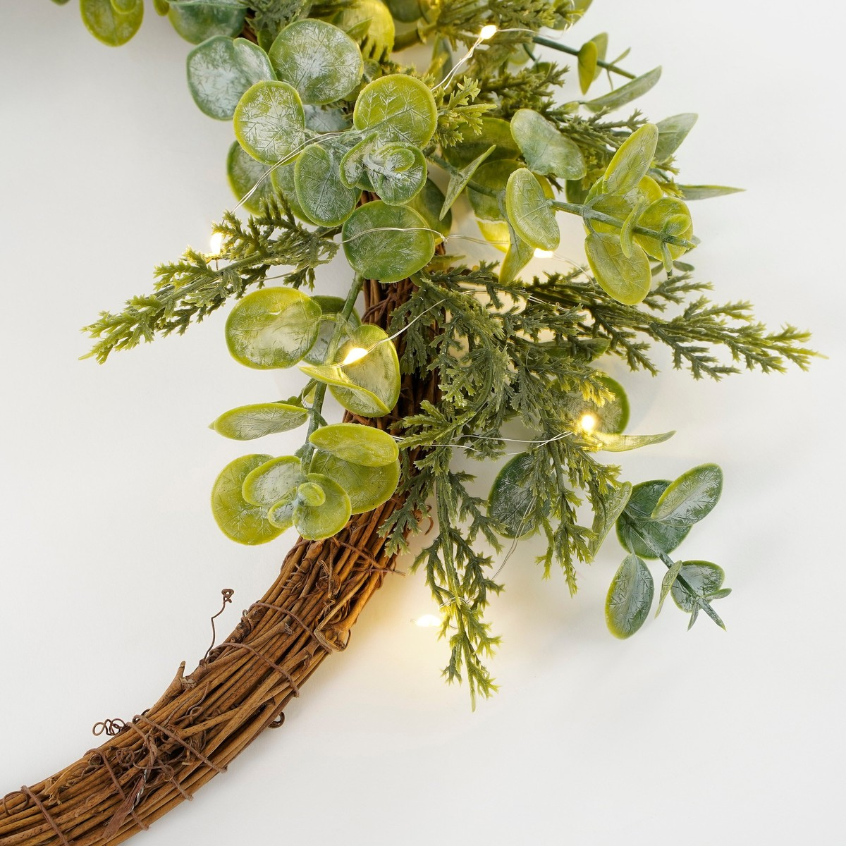 A twig wreath decorated with green eucalyptus and small warm white fairy lights on a white background.
