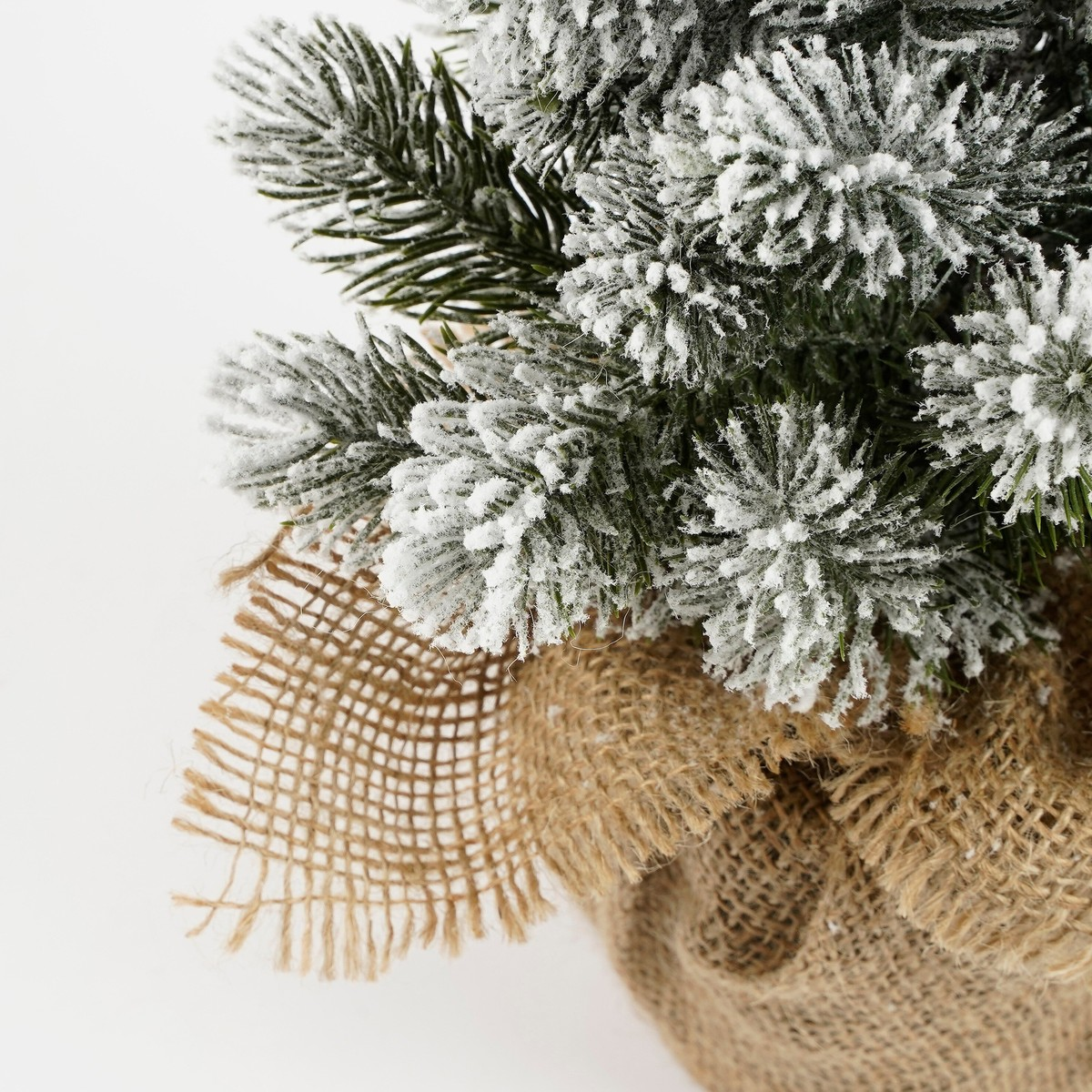 An artificial Christmas tree with faux snow on branches, wrapped in burlap, set against a white background.