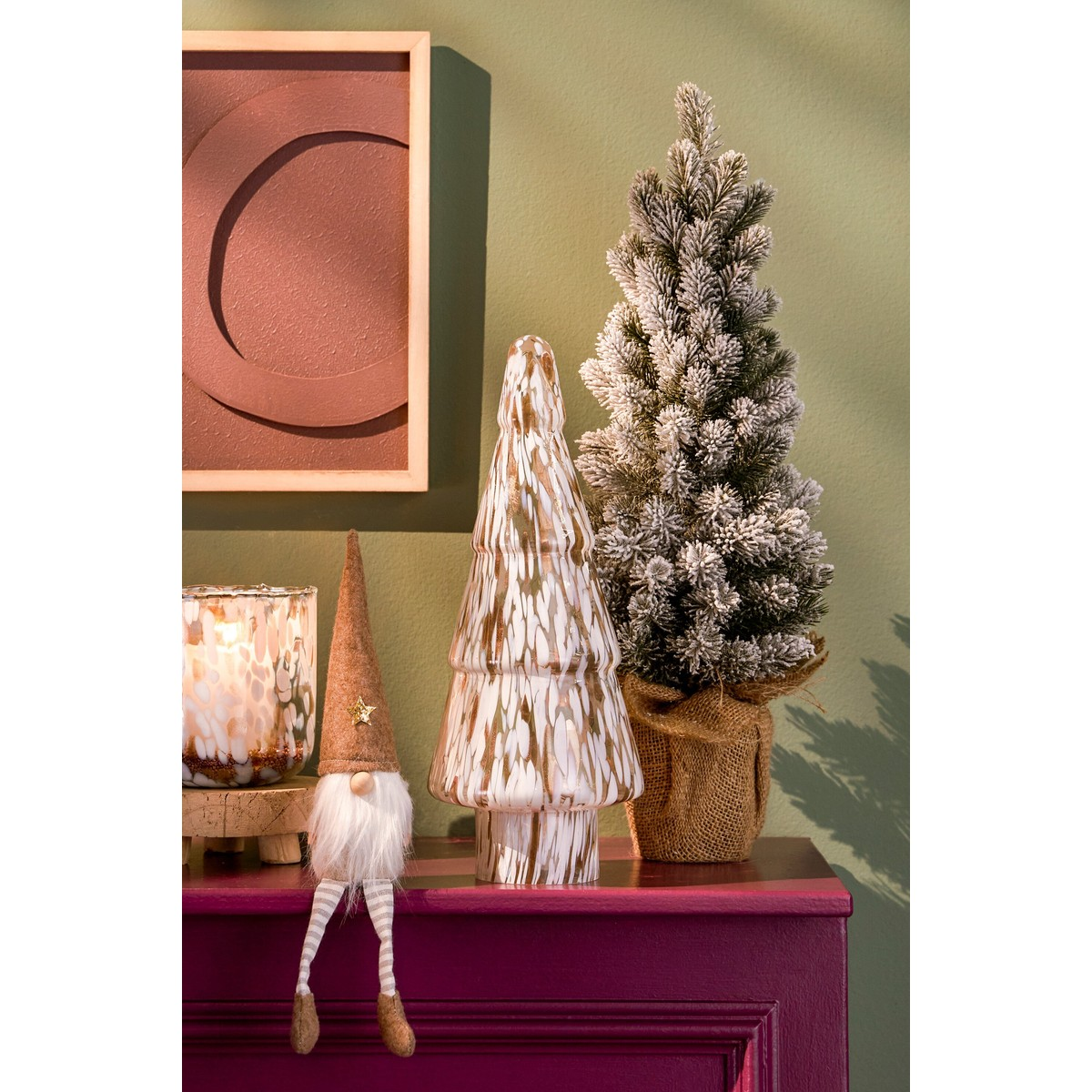 Holiday decor on a reddish cabinet with a gnome, glass tree, snow-flocked tree, and framed abstract art.