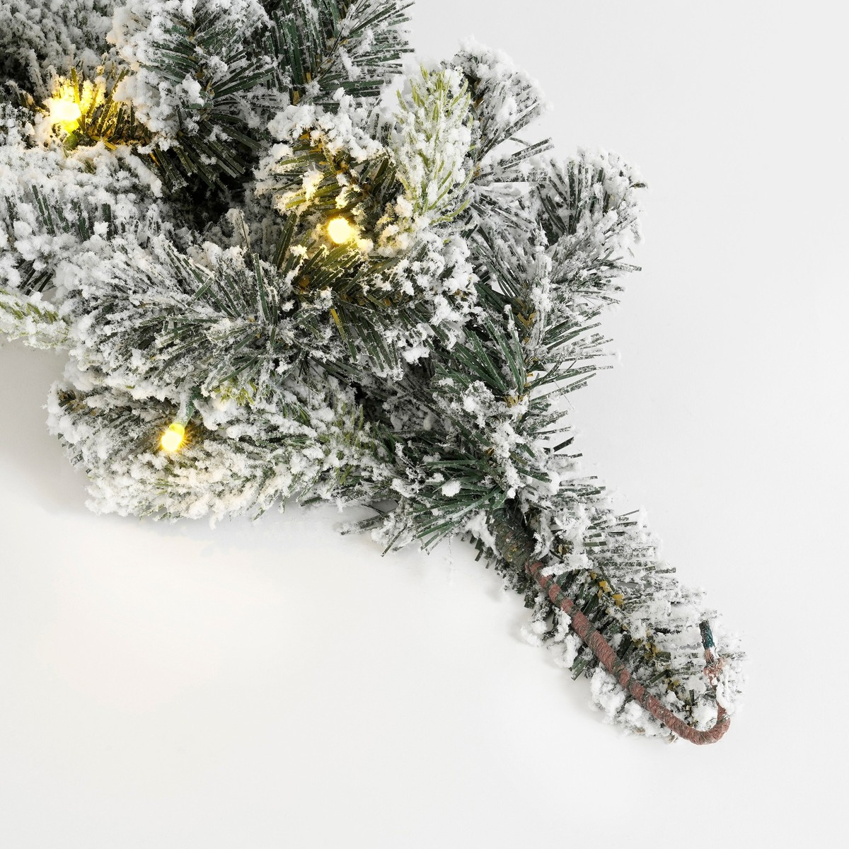 Verschneiter künstlicher Tannenbaumzweig mit warmweißen Lichterketten auf weißem Hintergrund.