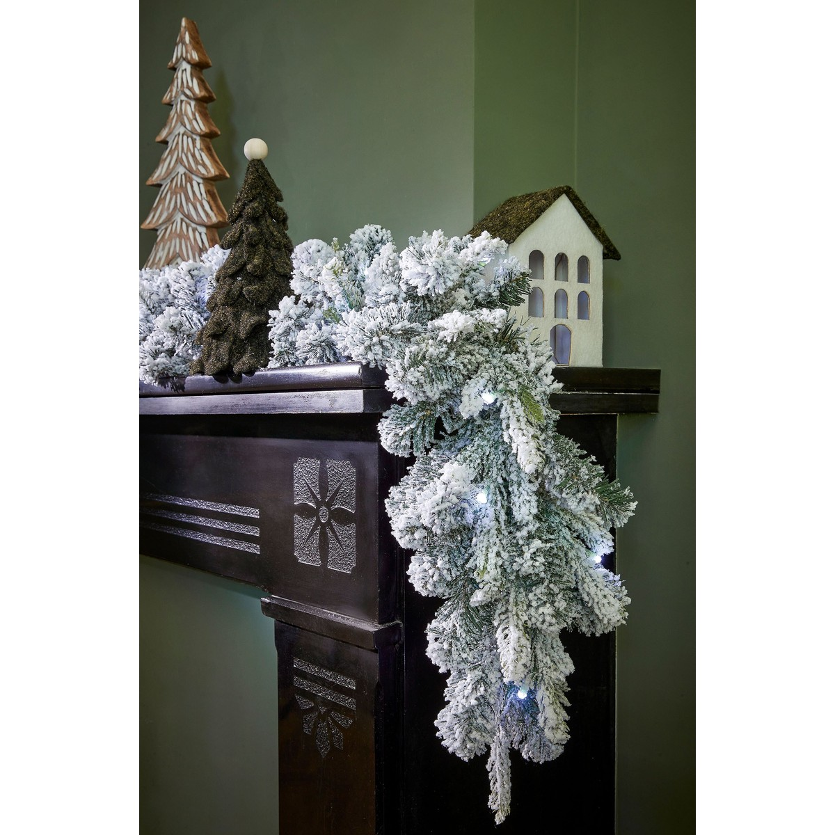 Ein schwarzer Kaminsims mit verschneiter Girlande, Holz-Weihnachtsbäumen und einem kleinen Haus auf grünem Hintergrund.
