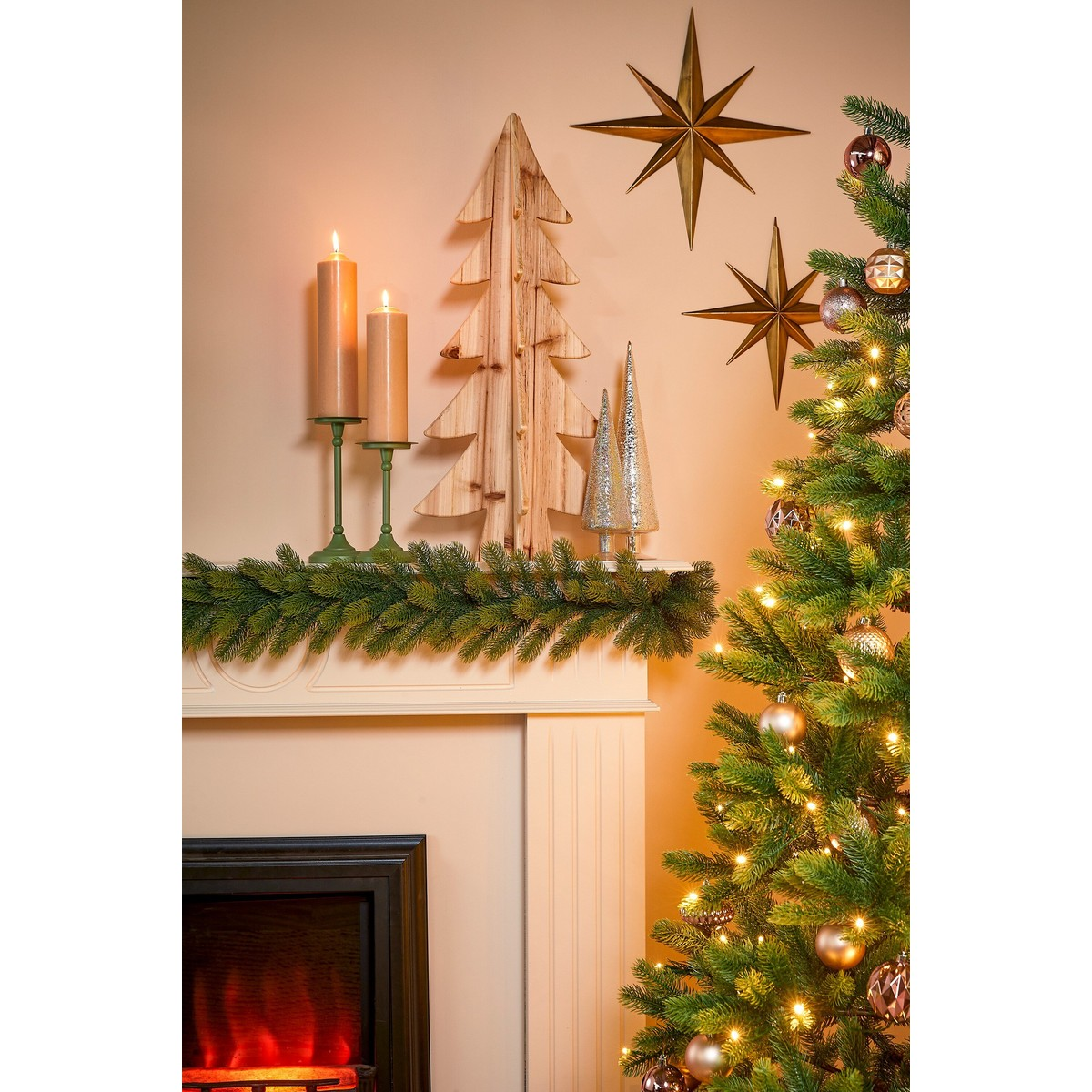 A decorated mantel with candles, wooden tree sculptures, and hanging stars beside a lit Christmas tree.