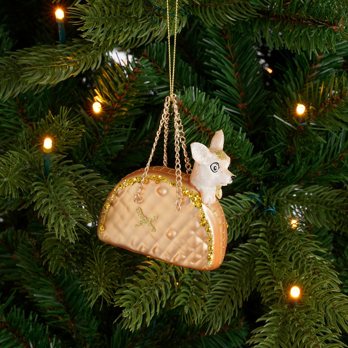 A chihuahua toy inside a gold taco ornament hanging on a lit Christmas tree.