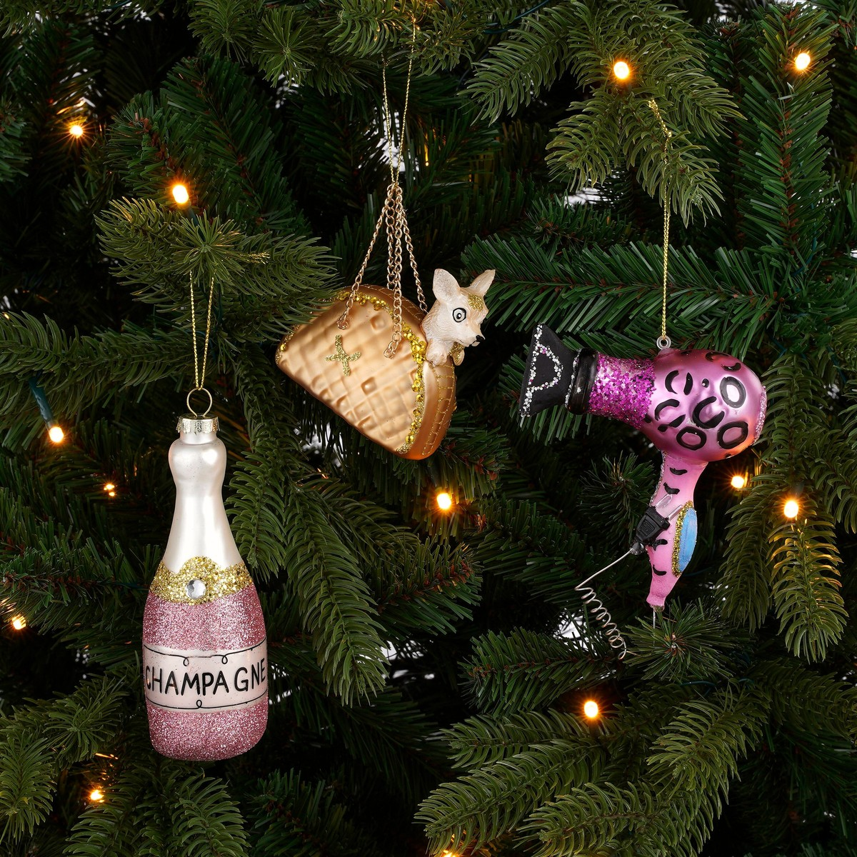 Christmas ornaments shaped like a champagne bottle, a dog in a handbag, and a hairdryer hanging on a green tree.