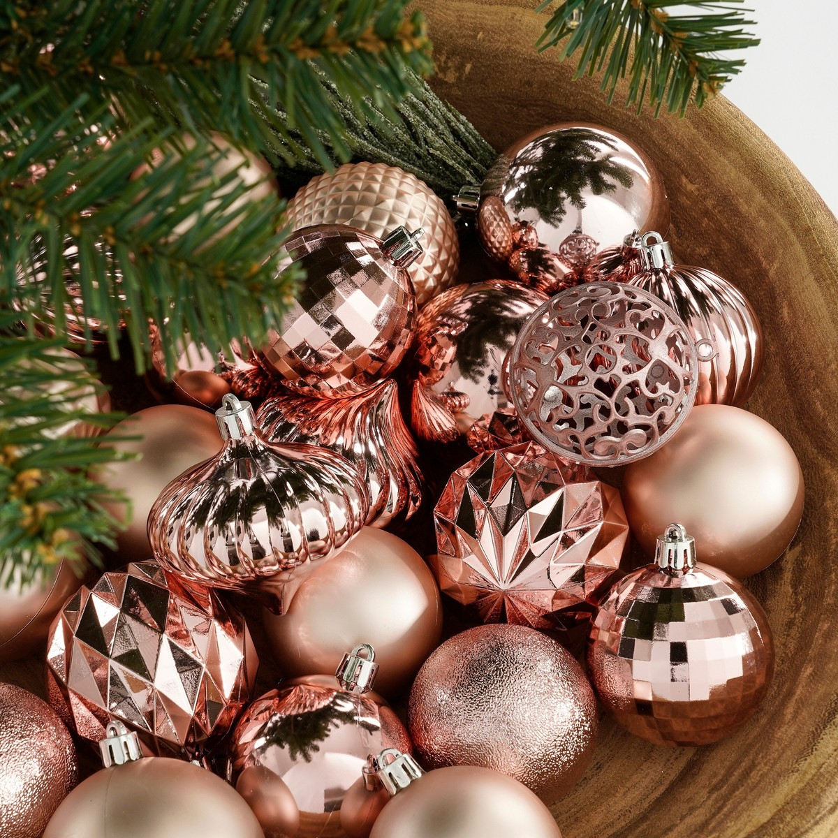 Shiny rose gold Christmas baubles in a wooden bowl with pine branches.
