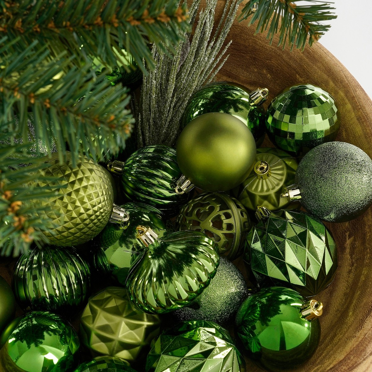 A wooden bowl filled with various green Christmas ornaments and pine tree branches.