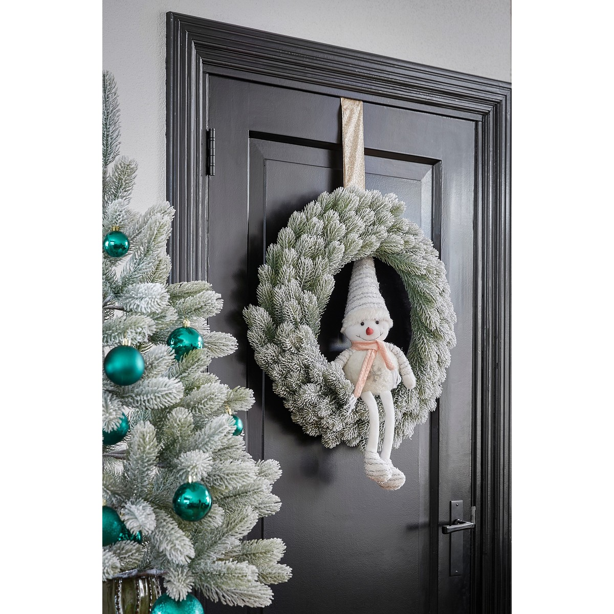 A snowy Christmas wreath with a plush doll hangs on a black door next to a decorated tree.
