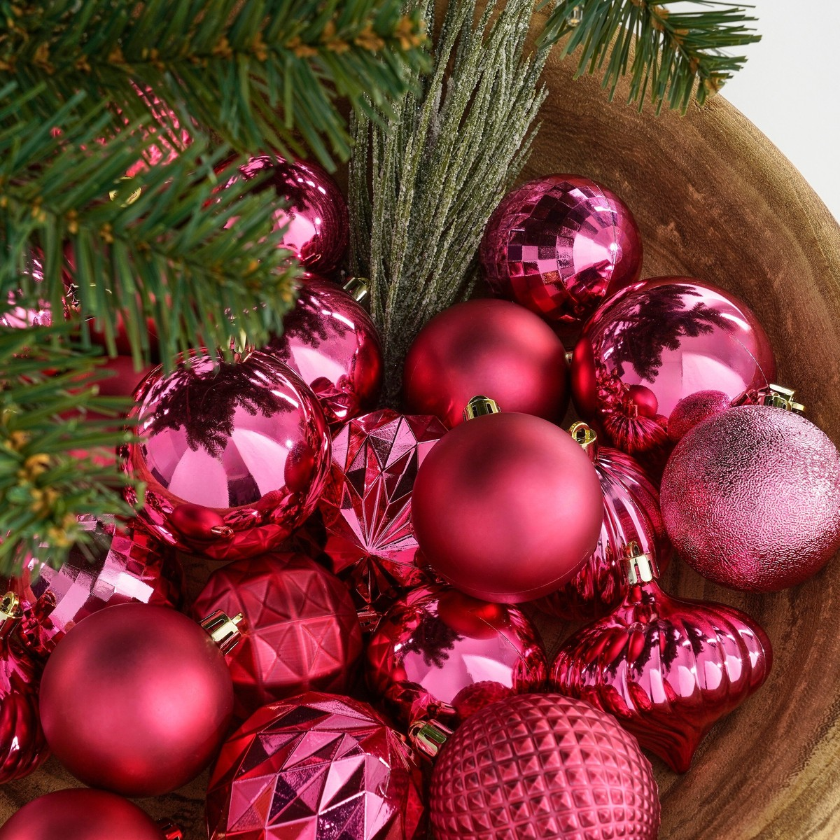 A wooden bowl filled with shiny pink Christmas ornaments and pine branches.