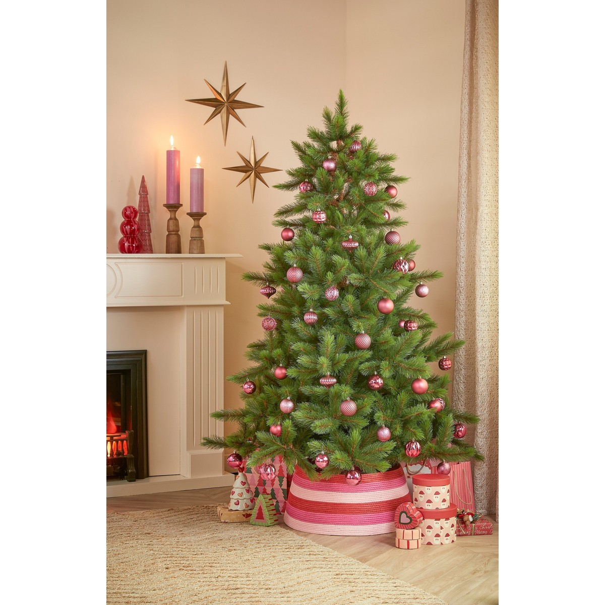 A Christmas tree decorated with pink ornaments stands in a cozy living room next to a fireplace.