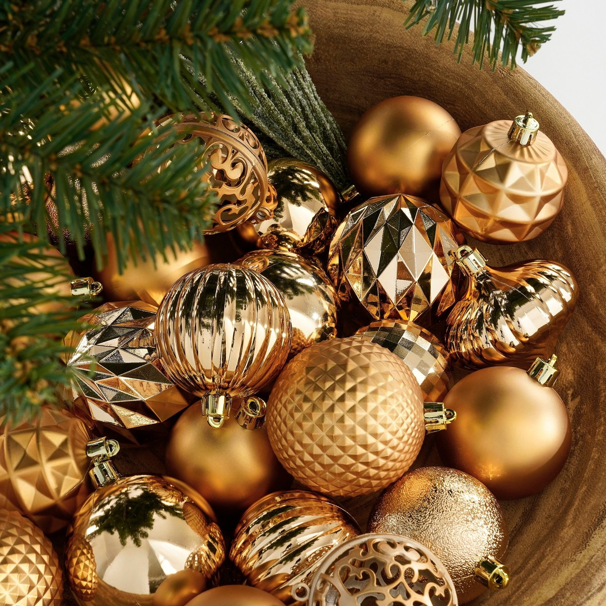 A wooden bowl filled with shiny gold Christmas ornaments and pine tree branches.