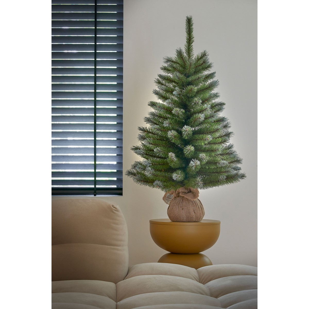 Small Christmas tree in burlap on a yellow table beside a sofa and dark window.