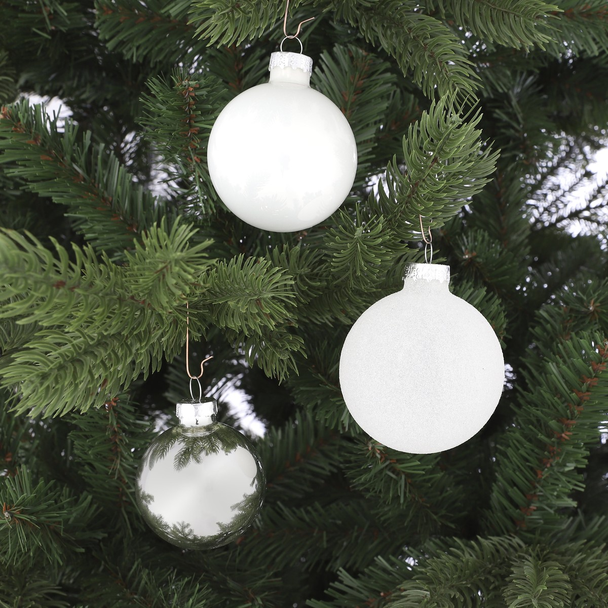 Three glossy Christmas baubles in white and silver hang from the green branches of an artificial Christmas tree.
