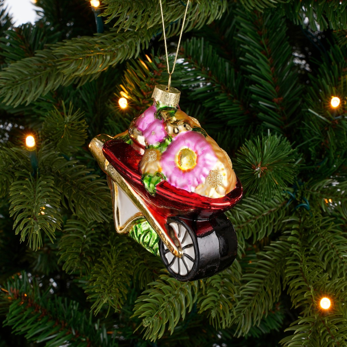 A colorful glass wheelbarrow ornament filled with flowers hangs on a lit Christmas tree.