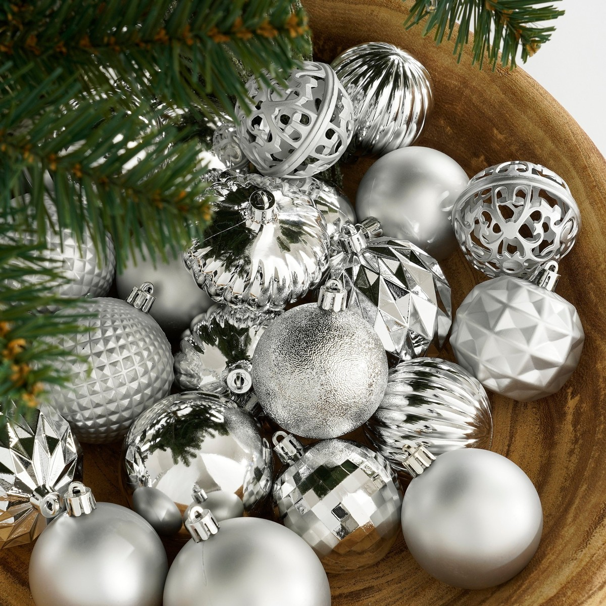 A wooden bowl filled with various silver Christmas ornaments and pine tree branches.