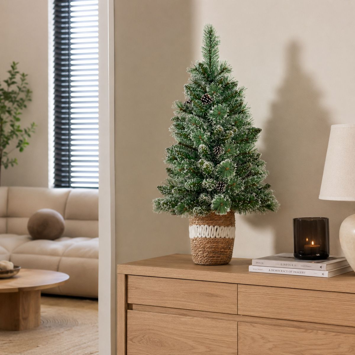 A small snow-dusted Christmas tree in a woven basket sits on a wooden cabinet, with a lit candle and books beside it.