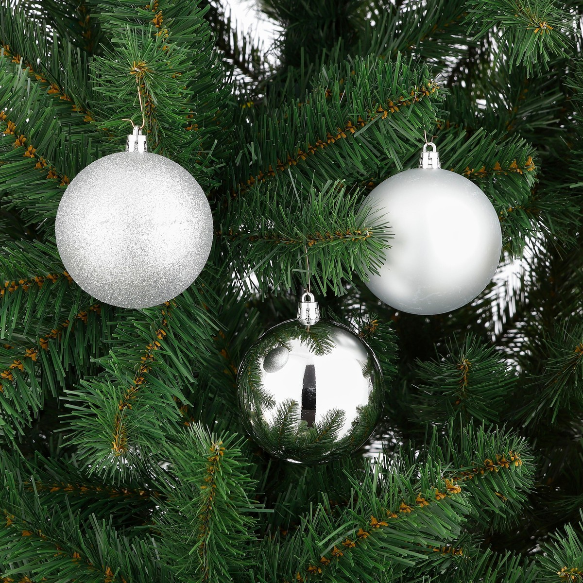 Three silver Christmas ornaments hang on a green artificial pine tree.
