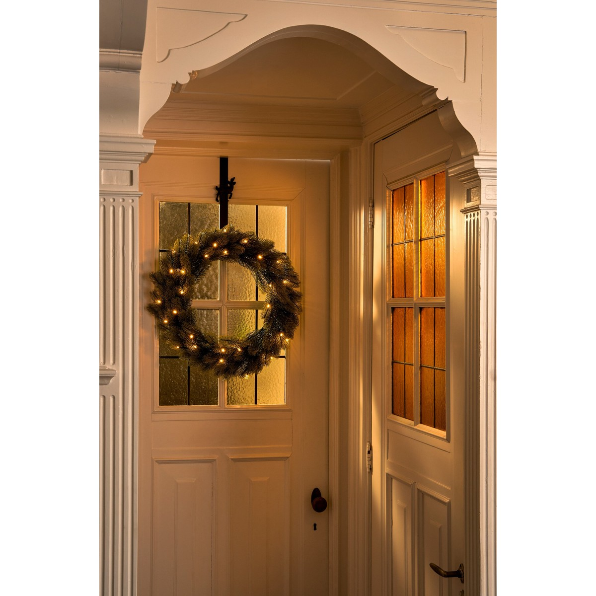 An illuminated Christmas wreath hangs on a white glass door in a hallway with warm lighting.