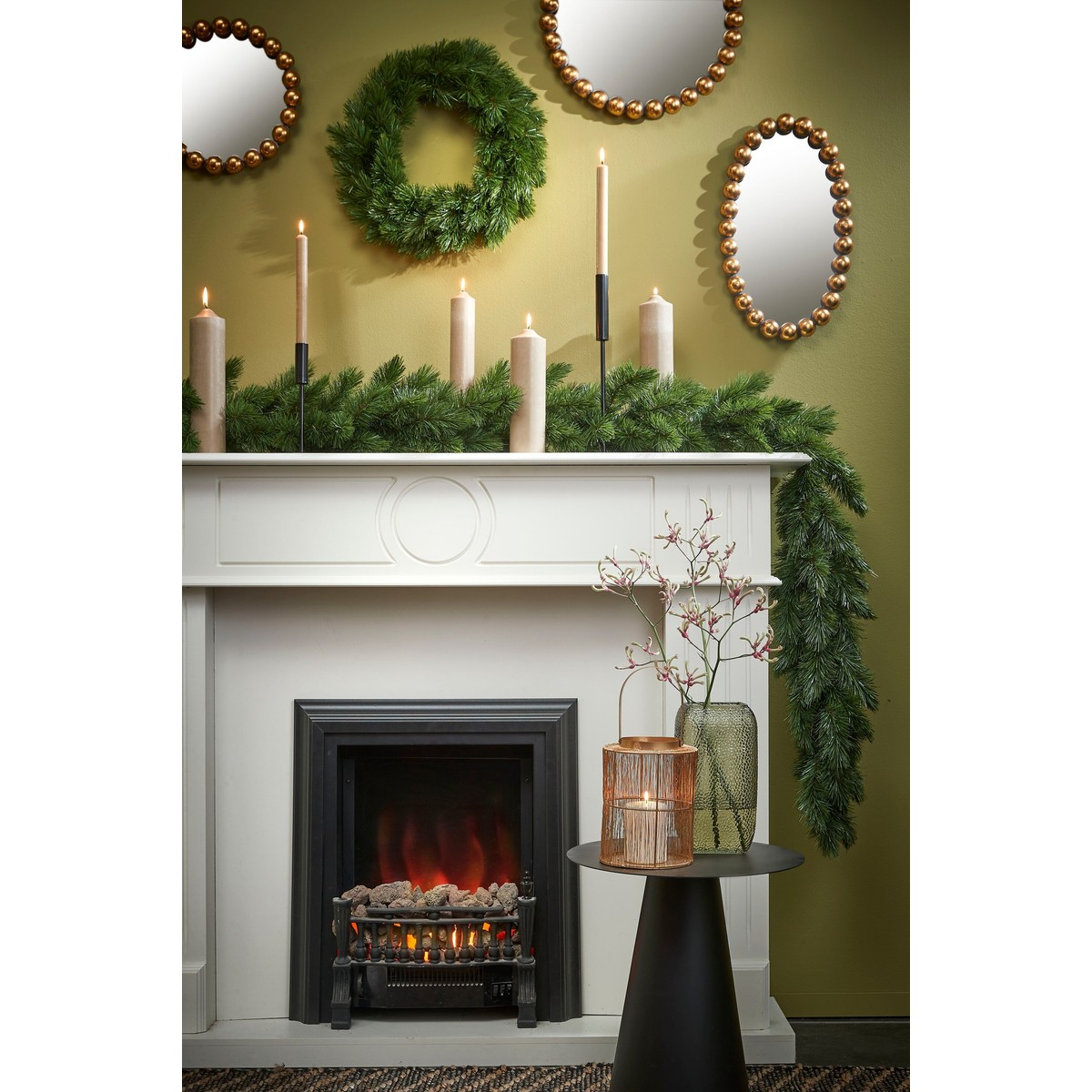 A white fireplace decorated with green garland, candles, mirrors, and a side table with vases.